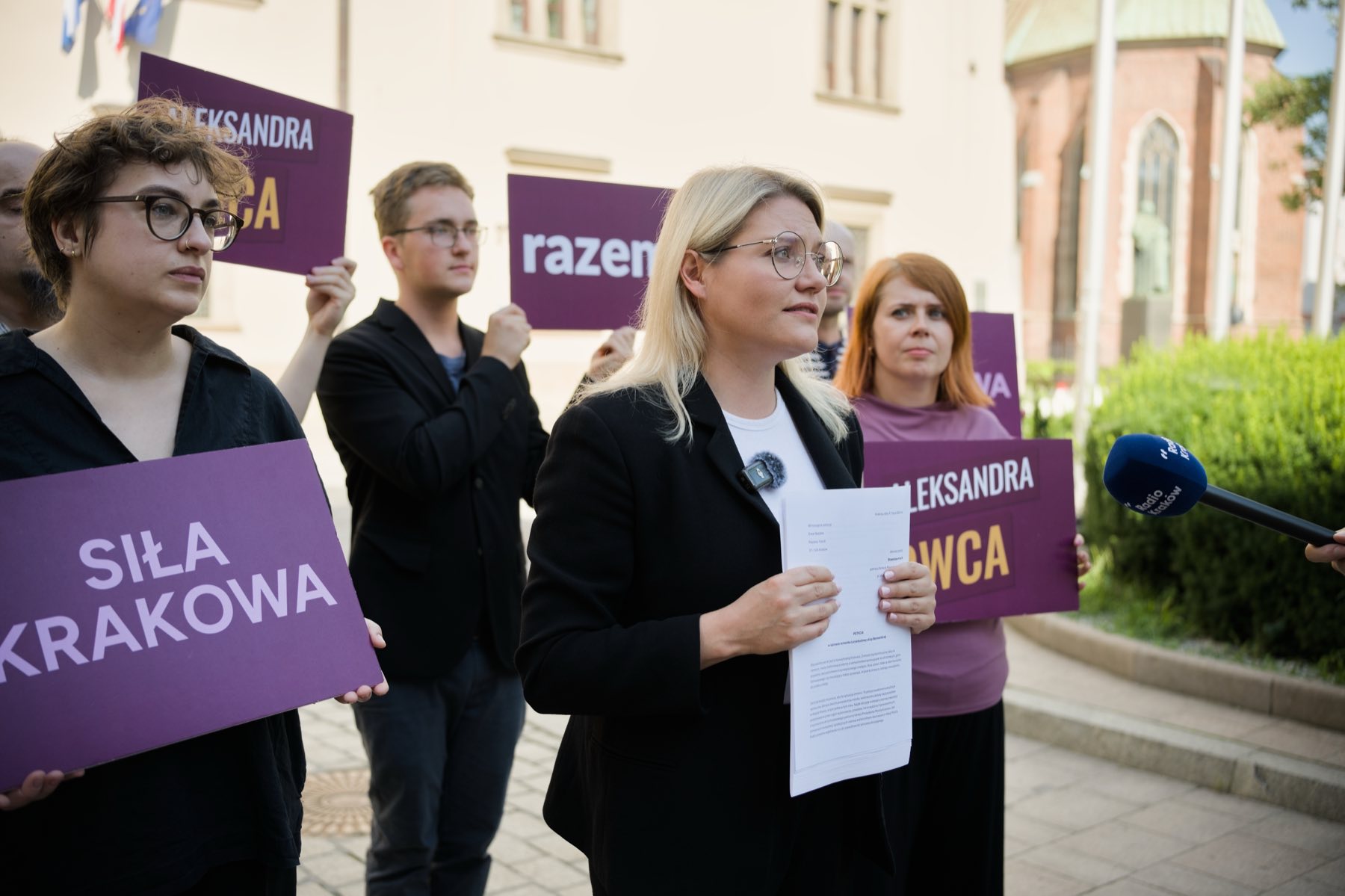 Aleksandra Owca podczas wystąpienia na konferencji prasowej przed Urzędem Miasta Krakowa, obok osoby trzymające tabliczki „Siła Krakowa” i „Aleksandra Owca”